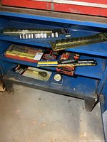 Assorted automotive tools on blue metal shelf including compression testers, Pocket Tuner, hydrometer, and socket sets in original packaging.