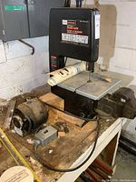 Side view of Craftsman band saw showing work table, saw blade, and motor setup on a wooden bench in a basement setting.
