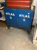 Front view of blue metal Atlas two-door cabinet showing brand name and overall condition.