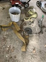 White bucket with various chains, heavy-duty yellow and beige straps, a manual cable winch, and black rolled strap all on a concrete floor.