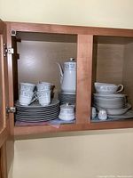 Photo of china set inside cabinet showing stacked plates, cups, saucers and a teapot with floral brocade pattern.