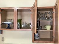Two open wooden cupboards with various glassware and mugs. Left cupboard contains blue pedestal cake plate, green pitcher, clear glass serving pieces. Right cupboard contains clear stemware on upper shelf, cream and yellow mugs, floral teacups with saucers on lower shelves.