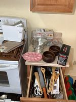 View of kitchen counter and stove corner with Hamilton Beach electric carving set box, Rival portable hand mixer box, two wooden bowls, pink ceramic egg tray, metal cake pan, and numerous kitchen utensils spread on countertop and drawer.
