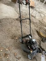 Full angle view of black Murray lawn mower with metal handle placed on dirt ground indoors.