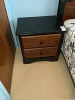 Nightstand beside bed showing black top and brown wooden drawers with black knobs, and size around 24x16x24 inches