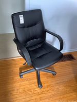 Front angled view of black vinyl office chair, showing cushioned seat, backrest, armrests, and 5-wheel base on wood flooring.