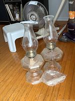 Pair of two clear glass oil lamps with octagonal bases and chimneys placed on wood floor, along with new oil lamp wicks in plastic.