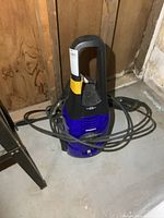Photo showing full blue and black Simoniz S1600 pressure washer with power cord wrapped around unit, placed on basement floor against wood wall.