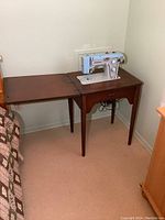 Wide view of vintage Kenmore sewing machine in wood cabinet with fold-out side extension next to bed.