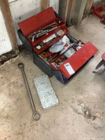 Photo showing vintage metal toolbox open with various tools inside, and large wrench and metal case outside on a concrete floor.