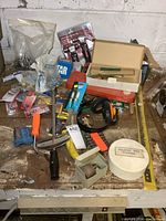 Overview photo showing assorted hand tools and hardware including wrenches, hammers, fasteners, and packaging items spread on a surface.