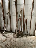 Wide view showing several long-handled yard tools leaning against wall, including multiple shovels, pitchforks, and a rake.