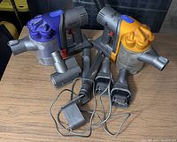 Two Dyson handheld vacuum cleaners with purple and yellow accents surrounded by accessories including multiple cleaning heads, extension wands, and chargers on a wooden surface.