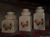 Three cream-colored stoneware sugar and spice containers with sunflower floral pattern on the front, set on a patterned cloth surface.