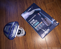 Front view of the blue Conair Steam & Press Turbo handheld garment steamer next to its retail box on a wood floor.