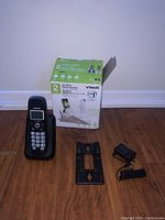 Cordless phone system components including 2 handsets, charging base, power adapter, wall mount and original box placed on wood floor against white wall