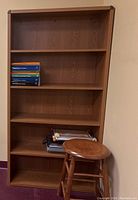 Wooden 5-shelf bookcase with multiple books stacked on the second shelf. Wooden round stool in front, not included.