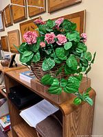 Artificial pink rose flower arrangement with green leaves in woven basket placed on wooden shelf.
