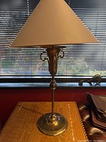 Full view of the brass table lamp on a surface in front of a window with blinds. The lamp has a conical beige shade and a brass body with a round base.