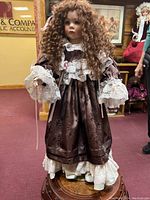 Front view of the porcelain jointed doll dressed in a brown fabric dress with white lace trim, standing on a stand.