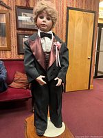 Front view of reproduction porcelain doll dressed in black tuxedo with burgundy satin lapels and boutonniere, standing on white base.