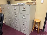 Photo shows four white metal filing cabinets in an office room against a wall. Two cabinets are locking, two are non-locking. Each cabinet has four drawers with handles and label holders.