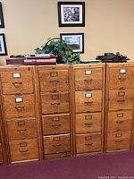 Photo of four wooden oak filing cabinets, each with four drawers, metal label holders and pull handles. Cabinets are arranged side by side against a wall with framed pictures and decoration on top.