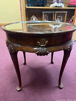 Front view of round wooden end table with glass top, brass handle, and carved leg details.