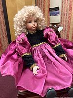 Front view of the porcelain doll seated, showing detailed face, curly blonde hair, pink satin puff sleeves, and black velvet dress with gold trim.