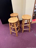 Photo showing set of three light oak wooden stools with round seats and four legs connected by stretchers. Wood grain and finish visible.