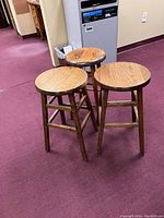Photo showing three wooden stools—two round and one square seat, all made from warm oak wood with visible wood grain and supports.