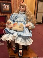 Front view of the porcelain sitting doll with long curly blonde hair, blue and white dress, holding a small stuffed bear, seated on a wooden chair.
