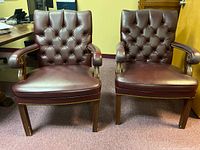 Front view of two burgundy vinyl office chairs with tufted backs and wooden legs and armrests, showing condition and style.