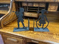 Photo shows two black wrought iron bookends placed on a wooden desk, one shaped as a boy playing a flute and the other as a girl skipping rope.