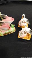 Photo of two pink ceramic fish figurines on a single base and two white stork figurines on individual bases against a black background.