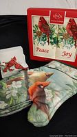 Photo showing two Lenox fingertip towels with cardinal and floral design, cardinal-themed oven mitt, and cardinal-themed coaster set with raised cardinal decoration