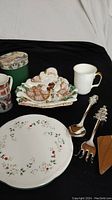 Photo showing the Santa dish with detailed Santa Claus design holding cookies, two Christmas mugs, the white mug with gold handle (Royal Albert), partial view of the holly patterned plate, and the silver plated utensils laid out on black fabric.