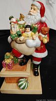 Front three-quarter view of Santa figurine holding a toy sack, standing on rooftop base with chimney and gifts.