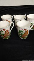 Five white ceramic mugs with robin bird and red berries motif, arranged on black surface.