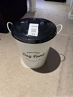 White metal dog food canister with black lid on carpeted floor, showing front label and handles.