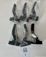 Five black guitar holders with padded arms still wrapped in plastic and hardware in small bags laid out on a white surface.