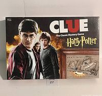 Front view of the Harry Potter Clue board game box showing main artwork of Harry, Hermione and Ron alongside a window showing part of the Hogwarts gameboard.