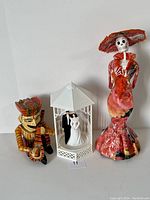 Lot containing all three figures: Mexican paper mache skeleton, wedding topper in gazebo, and Indian wooden figure placed side by side on white background.