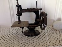Side view of the antique Singer toy sewing machine on a patterned surface showing the hand crank wheel and sewing platform.