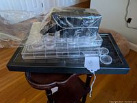 Clear acrylic coin holders with multiple round compartments, black coin display boxes in plastic wrap on top of a black tray, placed on wooden table and smaller wooden stool.