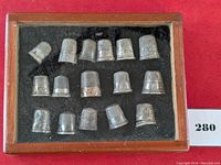 Display case holding 15 sterling silver thimbles arranged on black felt in wooden frame with glass top.