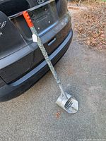 Full view of galvanized steel sand scoop with perforated scoop head and orange grip handle leaning against a car.