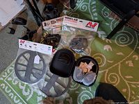 Overall shot on green rug showing two coil covers in packaging, a Minelab accessories bag, zippered headphone case and the sniper coil on a detector shaft.