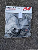 Sealed plastic package containing the black Minelab Equinox 11-inch coil metal detector accessory, with visible cables inside, on an outdoor paved surface.