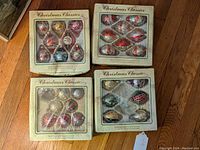 Four boxed sets of vintage Christmas glass ornaments placed on wooden floor, showing different shapes and patterns through plastic window panels.